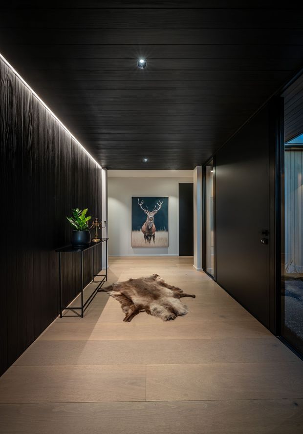 A dark, refined residential hallway with wide pale timber-look flooring, a near-black vertically textured feature wall and matching dark plank ceiling. Continuous linear LED lighting traces the wall-ceiling junction, while recessed downlights provide additional illumination. A slim black console with a potted plant and decorative objects sits against the wall, and a natural hide rug leads toward a softly lit end wall displaying a deer artwork. Dark internal doors and glazing line the right side.