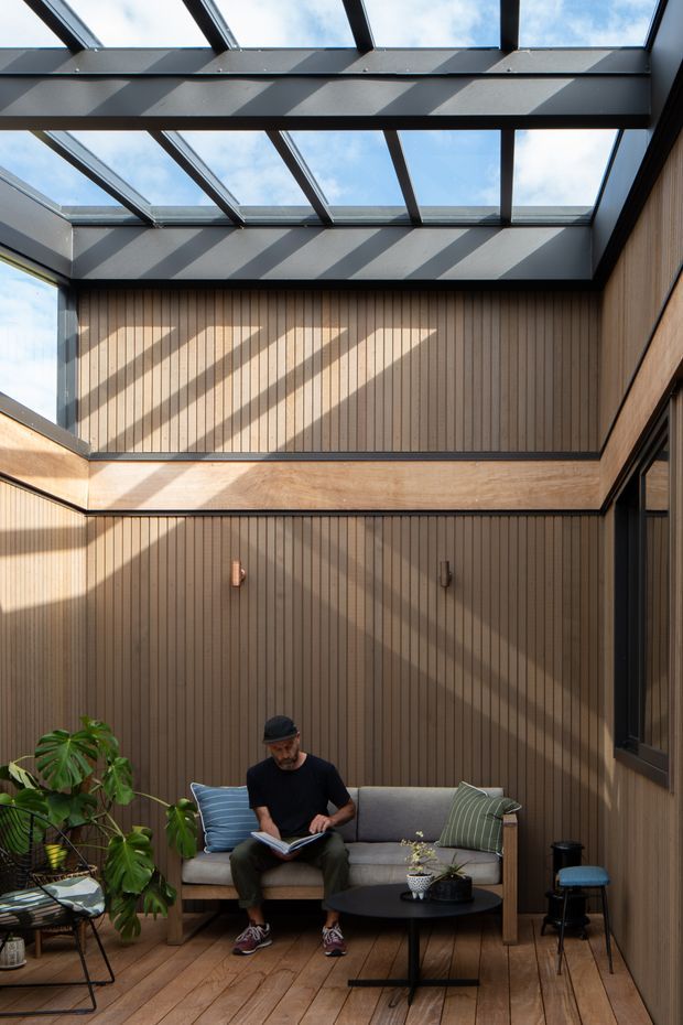A compact, warmly finished sunroom or enclosed outdoor living space with a dramatic glazed roof and clerestory-like skylight structure. Vertical timber wall panelling, timber flooring, dark metal framing, and large windows create a strong indoor-outdoor atmosphere. A small sofa with cushions, round coffee table, plants, and a person reading furnish the space. Sunlight casts bold diagonal shadows across the walls and floor.