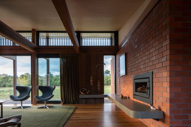 A warm mid-century-inspired living room with a prominent red brick fireplace wall, timber ceiling beams, dark timber wall panelling, and extensive floor-to-ceiling glazing overlooking a grassy landscape. Two sculptural lounge chairs sit on a muted green rug, while a long floating timber-toned hearth extends from the built-in fireplace.