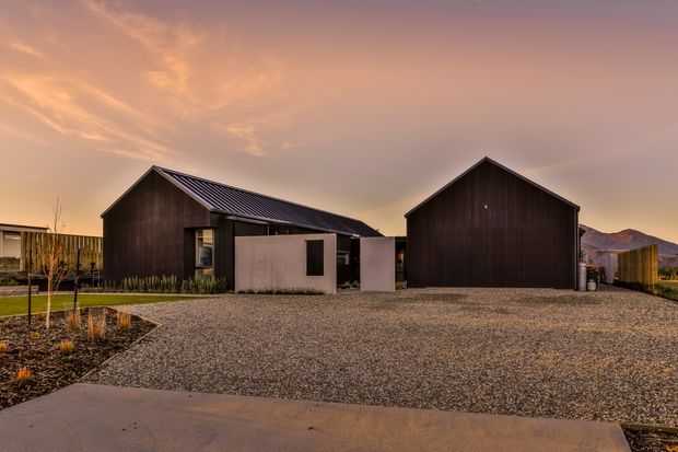 Exterior view of a contemporary property composed of two dark, gabled buildings with corrugated metal roofs and vertical dark cladding. A smaller central pale rendered volume connects the structures. The buildings sit around a broad gravel forecourt with minimal landscaped planting, viewed at sunset with mountains visible in the distance.