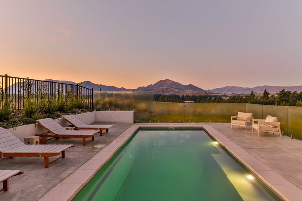 A contemporary rooftop or elevated outdoor pool terrace at sunset, with a rectangular swimming pool surrounded by pale paved decking, multiple timber sun loungers, and a small seating area. Transparent and metal privacy screens frame expansive views toward distant mountains and a softly coloured evening sky. The scene feels calm, refined, and resort-like.