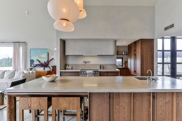 Open-plan contemporary kitchen with a large stone island and integrated sink, dark timber cabinetry, and timber island seating. A living area with a white sofa is visible to the left, while large windows and glazed doors bring in natural light. Sculptural pendant lights hang above the island, creating a warm, refined interior.