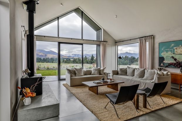 A bright contemporary living room with a dramatic exposed gable-shaped glazed wall and floor-to-ceiling windows overlooking a mountainous rural landscape. The room features a black wood-burning stove on a concrete hearth, polished concrete flooring, neutral sofas and lounge chairs, a low timber coffee table, woven rugs, sheer curtains, and a timber sideboard with artwork. The high sloping ceiling and expansive glazing create an airy, refined interior.
