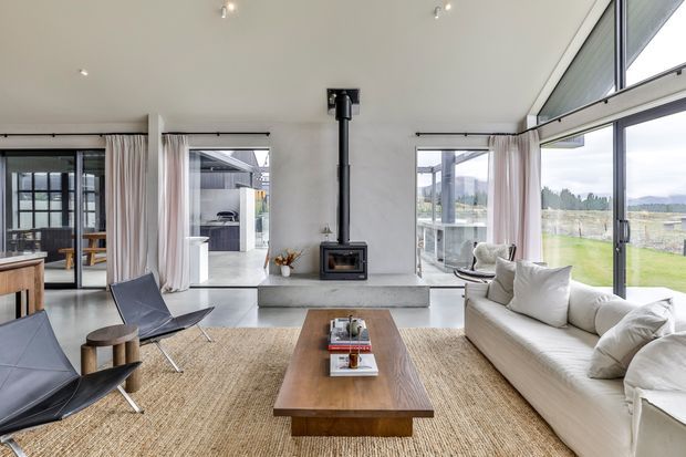 Bright contemporary living room with a high sloping ceiling, extensive floor-to-ceiling glazing, pale curtains, a white sofa, black leather lounge chairs, and a long timber coffee table over a natural woven rug. A freestanding black wood-burning stove is positioned on a raised concrete hearth, with views across an open rural landscape.