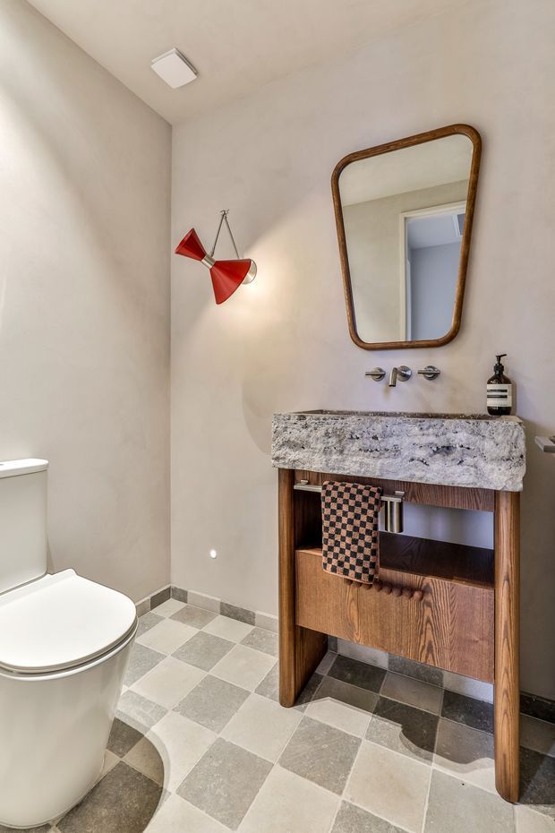 Compact powder room with a white toilet, wall-mounted basin, thick mottled stone benchtop, timber vanity cabinetry, checkerboard floor tiles, warm neutral walls, and a vintage-style framed mirror. A red articulated wall light provides a bold accent.