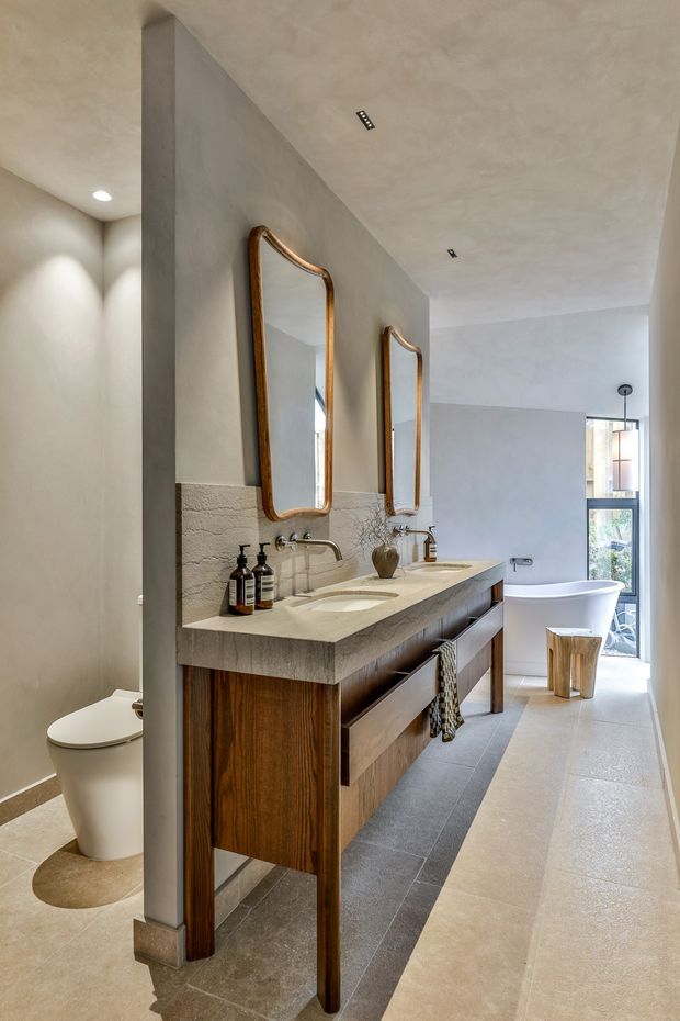 A refined contemporary bathroom featuring a long double vanity with a thick textured light stone benchtop, dark timber cabinetry, twin arched timber-framed mirrors, wall-mounted taps, and integrated basins. A freestanding bathtub sits near full-height glazing at the far end, while a partially screened toilet is visible to the left. The room has warm plastered walls, large pale stone or porcelain floor tiles, recessed ceiling lighting, and a calm neutral palette with natural timber accents.