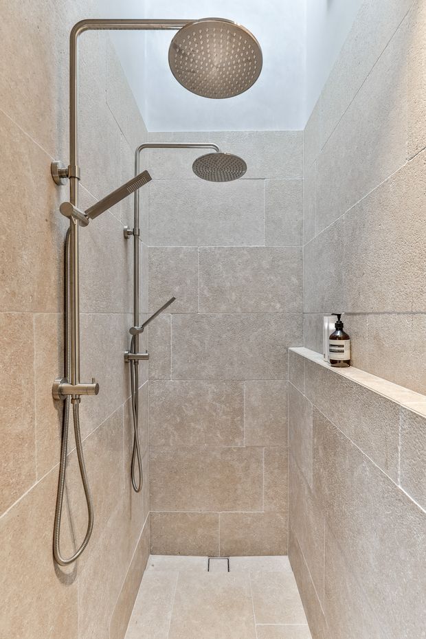 A compact walk-in shower enclosed by full-height beige-grey stone-look wall and floor tiles. The shower features dual rainfall showerheads, a handheld shower rail, brushed metal fixtures, a built-in tiled corner shelf with a soap dispenser, and a linear floor drain. The space is bright, clean, and minimalist with a neutral palette.