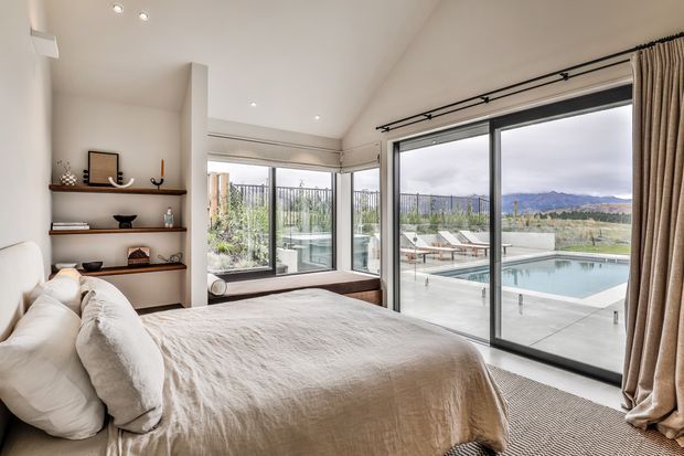 Bright contemporary bedroom with a large upholstered bed, neutral linen bedding, built-in open shelving, and expansive floor-to-ceiling glazing. Sliding glass doors open to a terrace overlooking a swimming pool and a broad rural or mountainous landscape. Recessed ceiling lights and simple timber shelving contribute to the clean, relaxed design.
