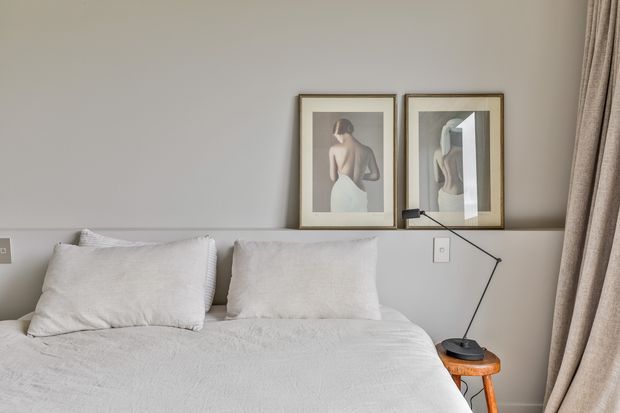 A softly lit, minimalist bedroom vignette featuring a neatly made bed with white textured bedding and pillows, a pale grey wall with a narrow ledge, two framed figure artworks, a small black angled desk lamp on a wooden bedside stool, and full-height beige curtains.