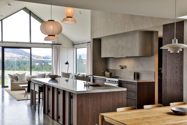 Open-plan contemporary kitchen and dining area with a large dark timber island, pale stone benchtops, integrated cooking appliances, and a timber dining table. The space connects to a bright living area through extensive black-framed glazing, with pendant lighting and mountain scenery visible outside.