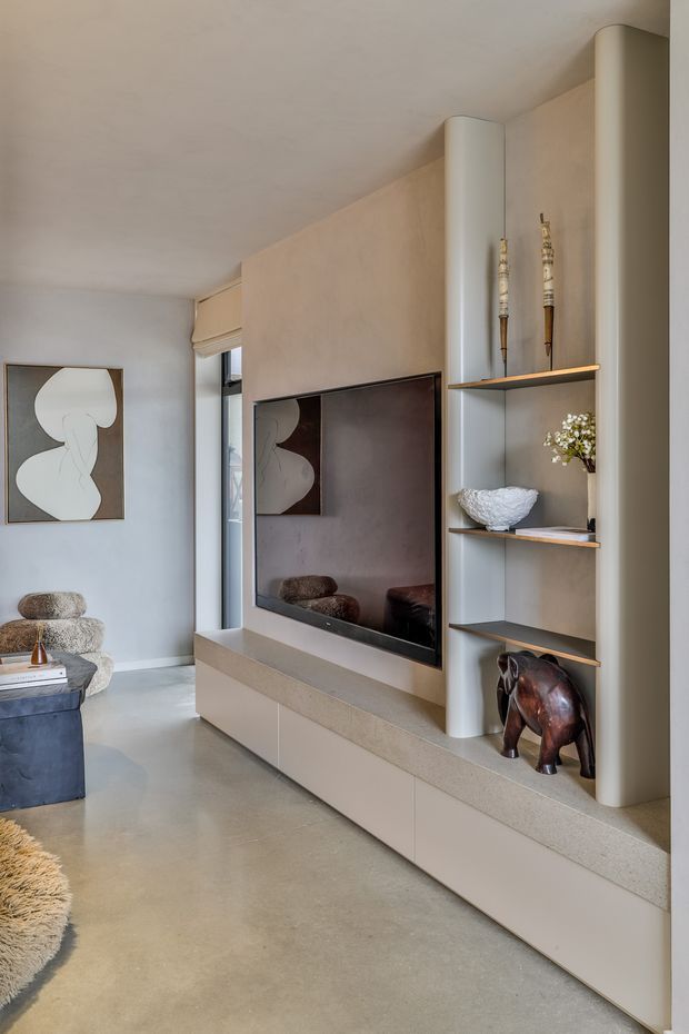 A refined contemporary living room featuring a full-height built-in media wall with a recessed television, pale taupe cabinetry, a stone-look ledge, open display shelving, and rounded vertical columns. The room has a smooth light concrete or polished screed floor, soft neutral walls, sculptural artwork, and understated decorative objects. A lounge chair and ottoman are visible at left, creating a calm, warm, minimalist atmosphere.