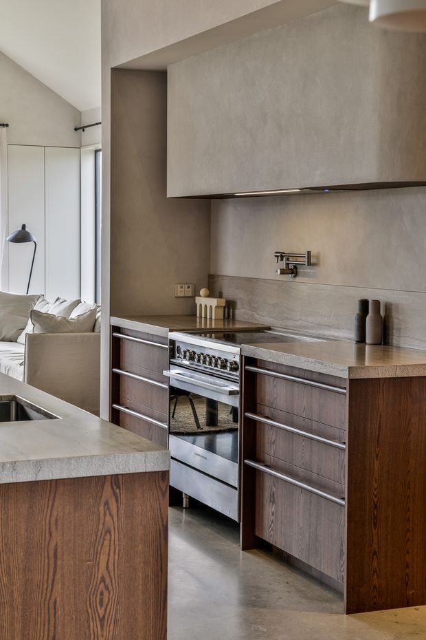 A refined contemporary kitchen with dark timber cabinetry, a large stone island, integrated stainless-steel range cooker, and a textured neutral plaster or concrete-style backsplash beneath a large concealed rangehood. The kitchen opens directly toward a softly furnished living area, with abundant natural light and slim black-framed glazing. The palette is warm and muted, combining brown timber, taupe-grey stone, and off-white walls.