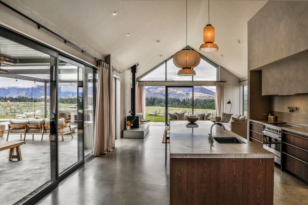 A refined open-plan kitchen and living space with a dramatic vaulted ceiling and expansive black-framed glazing overlooking a mountainous landscape. The kitchen features a large stone island with dark timber cabinetry, integrated cooking appliances and a sculptural pendant-light arrangement. Polished concrete flooring, neutral furnishings and warm timber accents create a contemporary alpine atmosphere. A glazed outdoor or sunroom area is visible to the left.