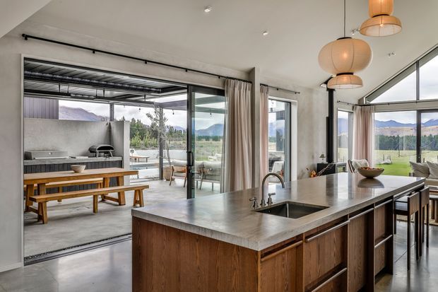 A spacious open-plan kitchen with a large timber island, dark stone benchtop, integrated sink and seating. Full-height glazing and sliding doors connect the kitchen to an outdoor entertaining area with a built-in barbecue and dining table, while pendant lights and a mountain landscape provide a warm, contemporary setting.