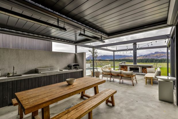 A large covered outdoor entertaining area with a built-in outdoor kitchen, dark vertical cabinetry, concrete benchtop, stainless-steel sink, barbecue and wood-fired oven. A timber picnic-style dining table sits in the foreground, while a relaxed outdoor lounge and fireplace occupy the open side. The space has a high exposed metal-lined roof with structural beams and pendant string lighting, opening onto a green rural landscape and mountains.