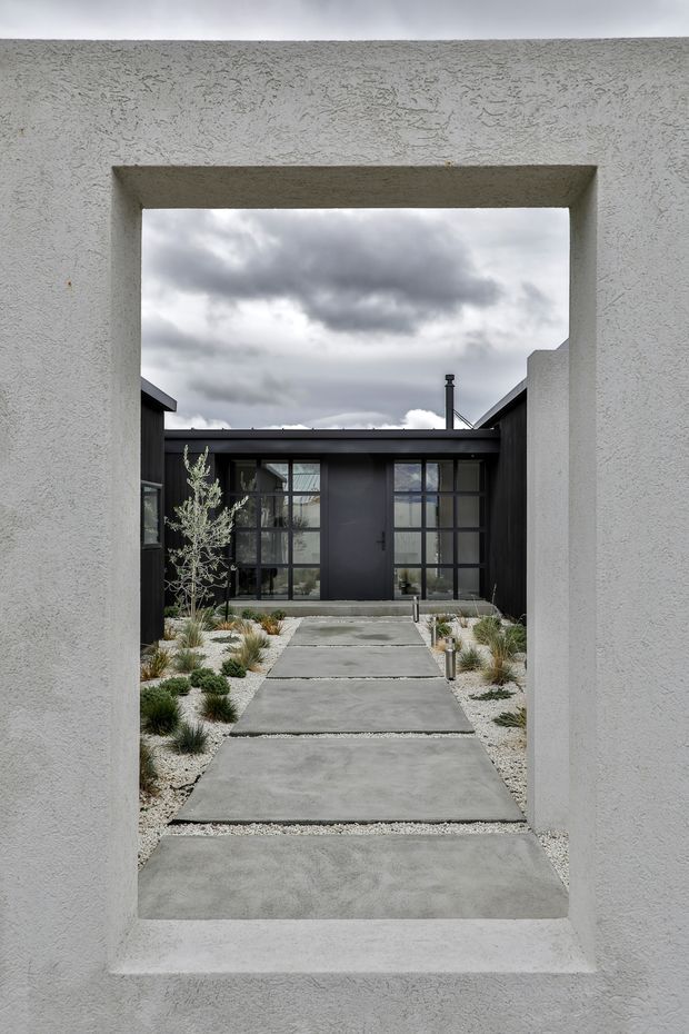 A minimalist courtyard entry viewed through a large rectangular opening in a textured white stucco wall. Stepping-stone concrete pavers lead through pale gravel and sparse ornamental planting toward a dark, glazed entrance facade with a central door and grid-framed windows. The composition has strong geometric lines and a moody overcast sky.