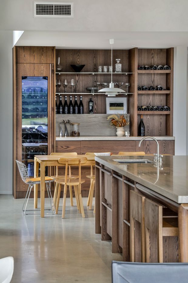 A refined open-plan kitchen and dining area with extensive dark timber cabinetry, integrated wine storage and glass-front wine fridge, a stone island with undermount sink, and a timber dining table with stools and wire-frame seating. Open shelving displays glassware and bottles, while a sculptural pendant light hangs above the island. The space has polished concrete flooring, a neutral palette, and a clean, contemporary built-in design.