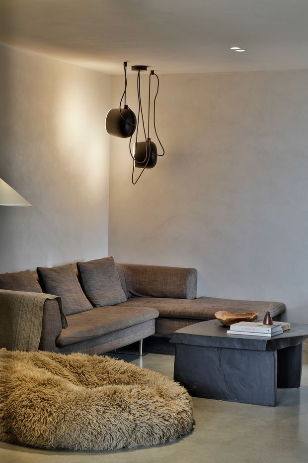 A warm, moody living room with a large taupe-grey sectional sofa, sculptural black pendant lights, a dark stone or concrete coffee table, and a shaggy tan rug. The room has softly textured plaster walls, a simple recessed ceiling, and minimal earthy decor.