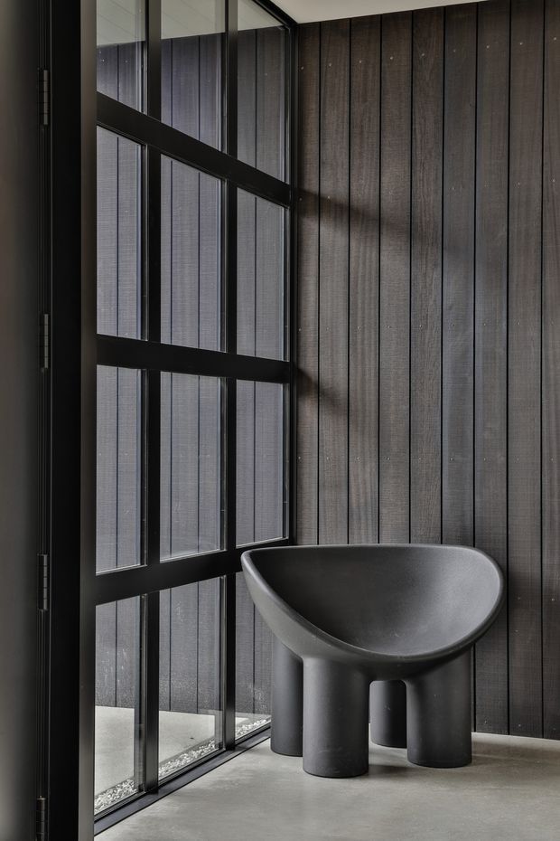 A dark, minimalist interior featuring a sculptural charcoal-grey freestanding basin or vessel sink positioned beside a full-height black-framed glazed wall. The adjacent wall is clad in vertical dark timber boards, while the floor is smooth grey concrete. Narrow blinds or screens filter light through the glazing, creating a moody, refined atmosphere with strong vertical lines and industrial detailing.