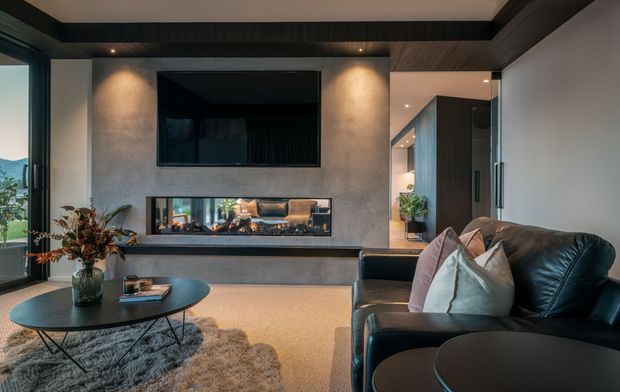 Contemporary living room with a black leather sofa, round coffee tables, shag rug and decorative vase arranged around a built-in fireplace. The fireplace wall has a textured grey finish, dark timber-look ceiling beams and a wall-mounted television. Full-height glazing opens to a green outdoor view, while a hallway and kitchen or utility cabinetry are visible beyond.