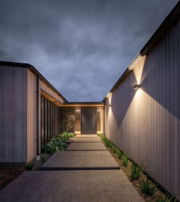 A moody dusk view along a narrow landscaped entry walkway between two modern single-storey building wings. The path is formed from large dark concrete slabs, bordered by gravel and low architectural planting, and leads to a glazed entrance door beneath a covered connection. Vertical dark cladding, warm timber-toned panels, concealed gutters, and wall-mounted uplights create a contemporary, refined exterior composition.