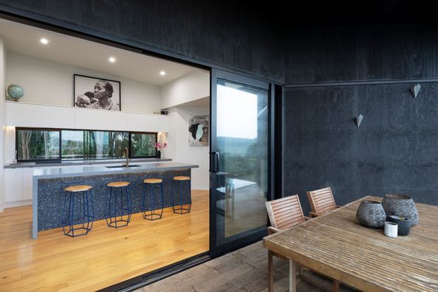 A contemporary open-plan kitchen and dining area viewed from an outdoor or adjoining terrace. The kitchen features a long island with a dark blue textured face, timber stools, white cabinetry, pale timber flooring, and a slim horizontal window. Large black-framed glazing separates the spaces, while the dining area has a timber table and chairs against a dark textured feature wall. A framed monochrome artwork and subtle decorative objects add visual interest.