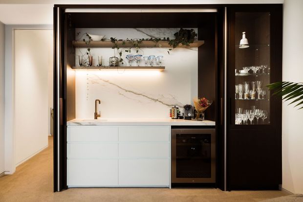 Built-in home bar and drinks cabinet set within a dark timber-framed alcove. The space includes white handleless cabinetry, a pale marble-look splashback, brass tap, open illuminated shelving with glassware and greenery, and a tall glazed display cabinet. Warm integrated lighting contrasts with the dark joinery and light carpeted surroundings.