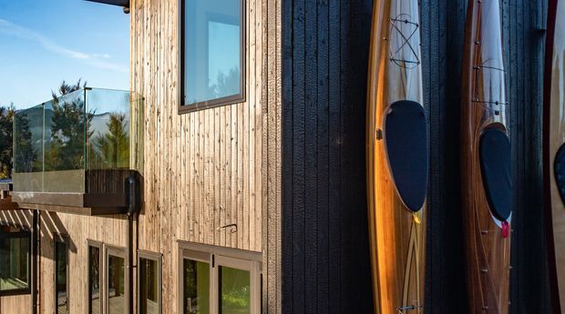 Close exterior view of a contemporary multi-level building clad in vertical natural timber and dark textured cladding. Large glazed doors and windows open toward a glass-balustraded balcony, while several surfboards are stored vertically against the facade. Trees and blue sky are visible in the background.