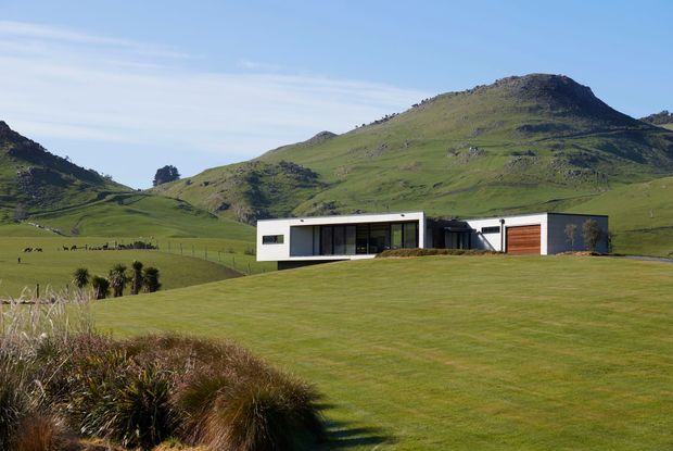 A low, modern single-storey residence set in an expansive green rural landscape, with a long rectangular white facade, dark floor-to-ceiling glazing, a projecting flat roof, and an attached garage with a timber-toned door. Rolling grass-covered hills dominate the background, while native tussock planting frames the foreground.