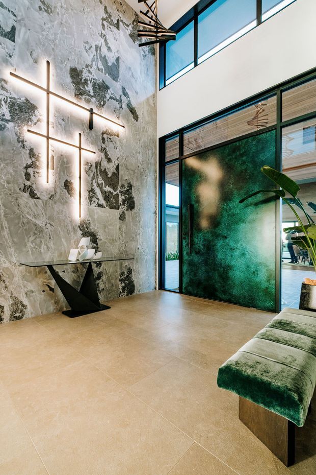 A dramatic contemporary entry hall with a very high ceiling, expansive black-framed glazing and an open doorway leading into adjoining spaces. The main wall is clad in heavily veined grey-and-white stone, complemented by sculptural black metal wall lighting, a slim glass console with a geometric black base, and a deep green upholstered bench. Warm beige flooring and natural light balance the bold, luxurious finishes.