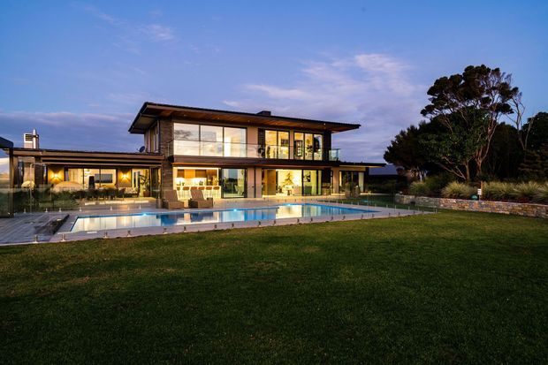Twilight exterior view of a large contemporary multi-storey residence with extensive floor-to-ceiling glazing, projecting flat roof planes, timber-toned cladding and illuminated interiors. A long swimming pool and paved terrace sit directly in front of the house, with broad lawn, planted borders and mature trees surrounding the property.