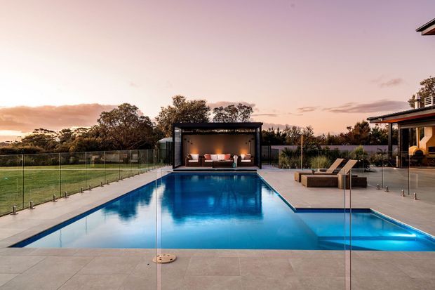 A contemporary outdoor pool area at sunset, viewed across a large rectangular swimming pool with a shallow sun shelf and clear glass safety fencing. A covered poolside pavilion with built-in seating and warm lighting sits at the far end, while loungers and a modern house are visible to the right. The setting includes landscaped trees and an expansive lawn beyond the property.