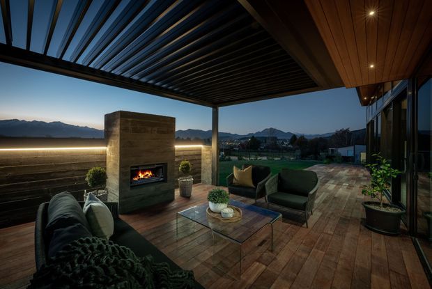 A covered outdoor terrace at dusk with a lounge seating arrangement, glass coffee table, and built-in outdoor fireplace. Timber decking, a timber-lined ceiling, dark overhead slatted screening, and extensive glazing frame views toward a mountain landscape. Warm integrated lighting creates an inviting evening atmosphere.