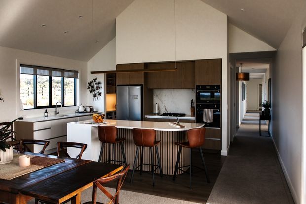 Open-plan kitchen and dining area in a contemporary home. The kitchen features a large island with a white stone-look benchtop, dark timber cabinetry, integrated appliances, a sculptural white volume above the cooktop, and warm pendant lighting. A dining table with leather chairs occupies the foreground, while a carpeted hallway extends toward the rear. Large windows provide natural light and views of the landscape.