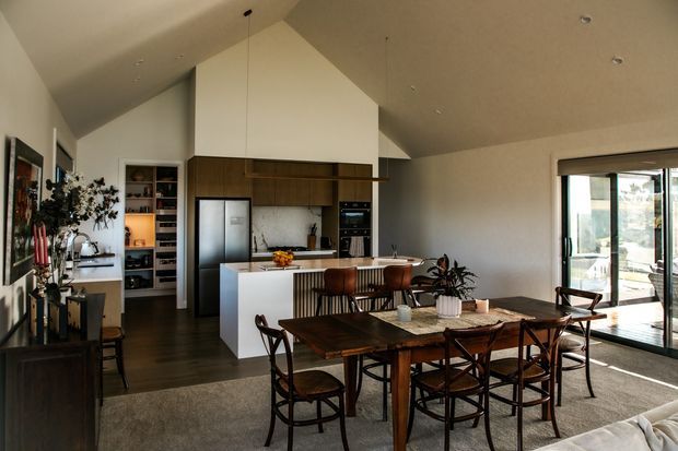 Open-plan contemporary kitchen, dining and living area beneath a high angled ceiling. The kitchen features dark timber cabinetry, a white island and integrated appliances, while a timber dining table with chairs occupies the foreground. Full-height sliding glass doors open to an outdoor area, and built-in shelving and warm wood flooring add texture to the neutral interior.