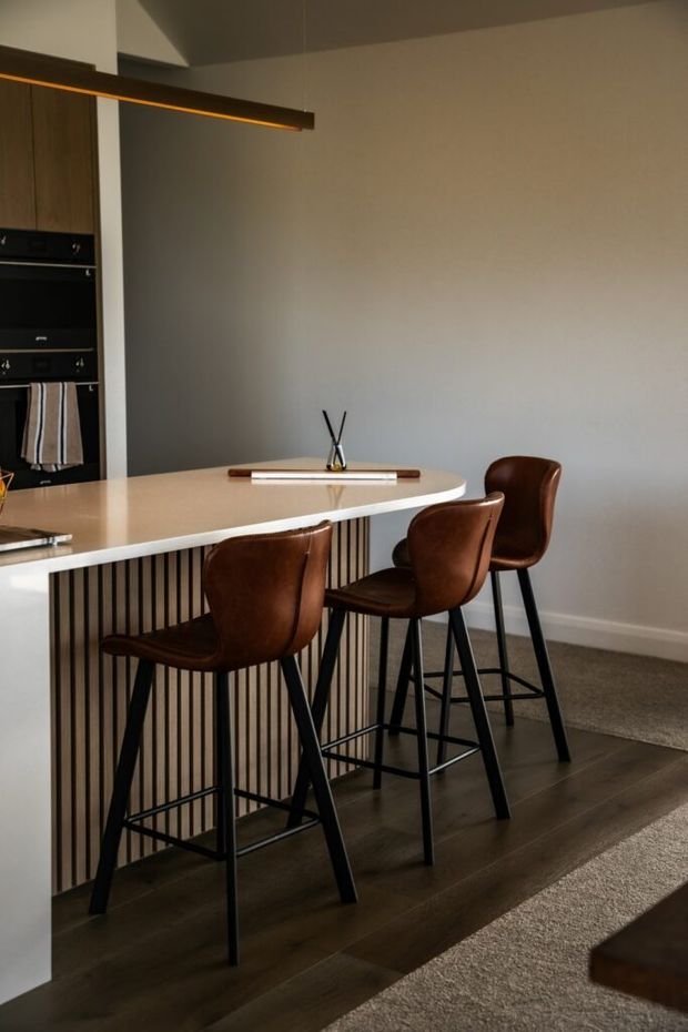 A contemporary open-plan kitchen and dining area with a curved pale stone island or breakfast bar, dark vertical slatted detailing, integrated black appliances and dark timber cabinetry. Three brown leather bar stools with black metal frames line the island. A slim linear pendant light hangs above, while warm dark timber flooring transitions to carpet in the adjacent space. The room has a restrained neutral palette and clean, refined detailing.