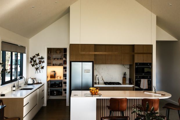 Contemporary kitchen with a dramatic vaulted ceiling, white walls, dark timber cabinetry, marble-look splashback, and a large island with integrated seating. The space includes a stainless-steel refrigerator, built-in black appliances, open shelving, extensive glazing, and warm timber flooring.