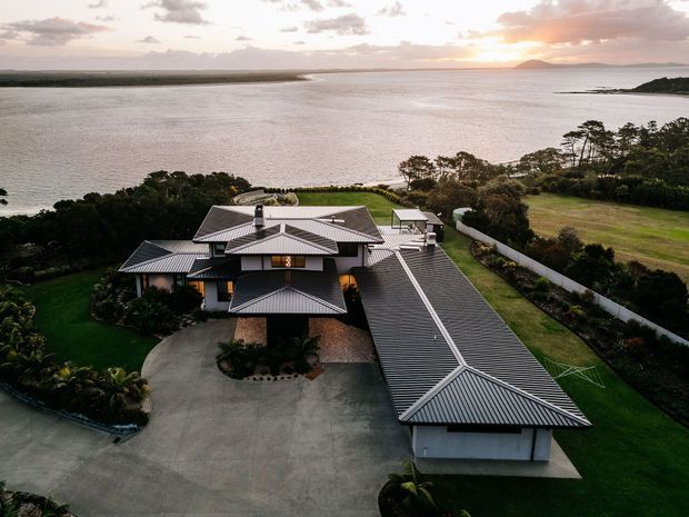 Aerial view of a large waterfront residence with multiple interconnected pitched metal roof forms, landscaped grounds, curved driveway and detached-looking covered wing. The property overlooks a broad calm body of water with a low, green shoreline and sunset beyond.