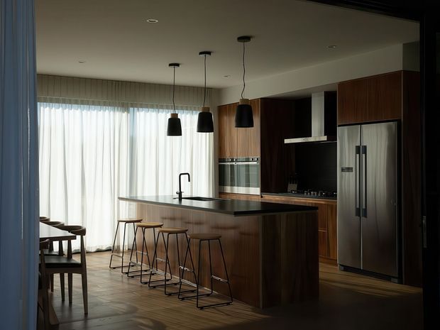 A sophisticated open-plan kitchen and dining area with dark timber cabinetry, integrated stainless-steel appliances, a large island with a dark benchtop, and a row of slim black pendant lights. Sheer floor-to-ceiling curtains provide soft daylight, while timber flooring and dining furniture create a warm, refined atmosphere.