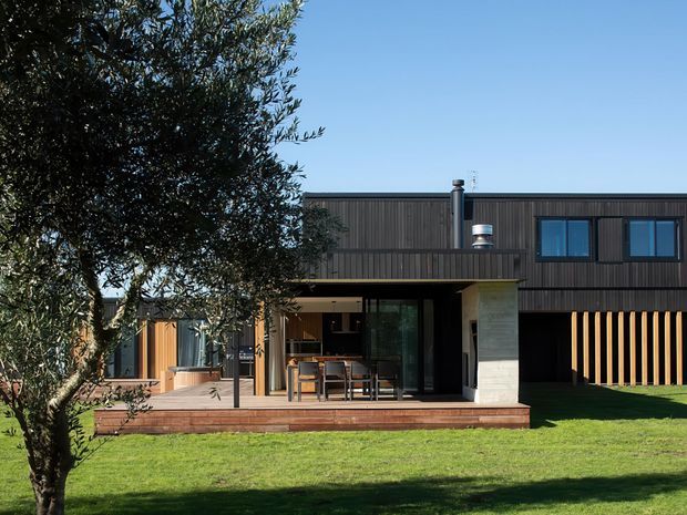 A modern two-storey house viewed from the rear garden, with dark vertical cladding, large glazed openings, a covered outdoor dining terrace, timber decking, and a prominent light stone or concrete wall element. Vertical timber screening defines the right side of the facade, while a broad lawn and mature tree occupy the foreground.