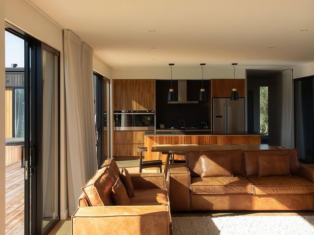 Warm contemporary open-plan living room and kitchen with extensive dark-framed glazing, timber cabinetry, a black island and backsplash, pendant lighting, and substantial tan leather seating. A timber deck is visible through the glazing, while sunlight creates a bright, inviting atmosphere.