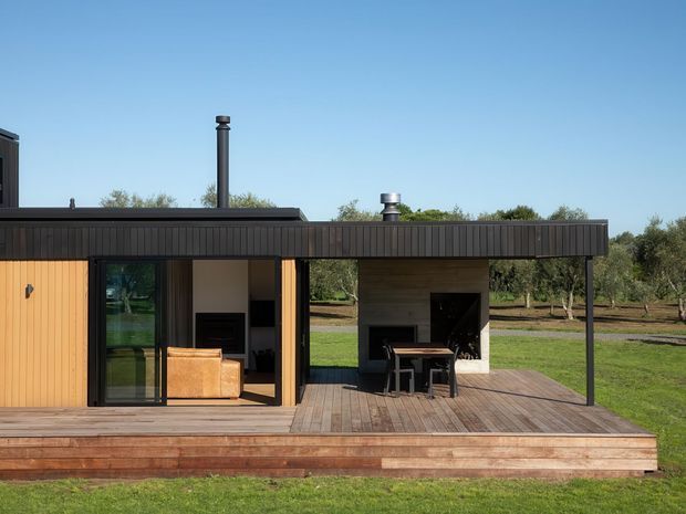 Single-storey modern pavilion-style home viewed from the garden, with a low flat roof, dark vertical cladding, warm timber wall panels, extensive glazed openings, and a broad timber deck. The covered outdoor area contains a dining table and built-in fireplace or barbecue zone, while a living area is visible through the glazing. The house sits in a landscaped rural setting with established trees and lawn.