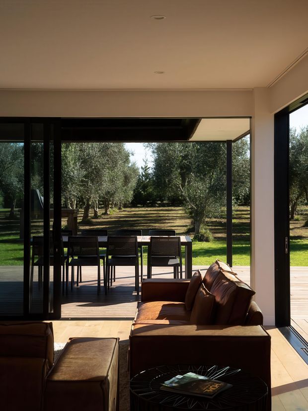 A warm, modern living room opens through large black-framed sliding glass doors to a timber deck and outdoor dining area, with an olive grove beyond. The interior contains a brown leather sofa and ottoman, a dark wire-frame coffee table, recessed ceiling lights, pale walls, and timber flooring. The expansive glazing creates a strong indoor–outdoor connection.