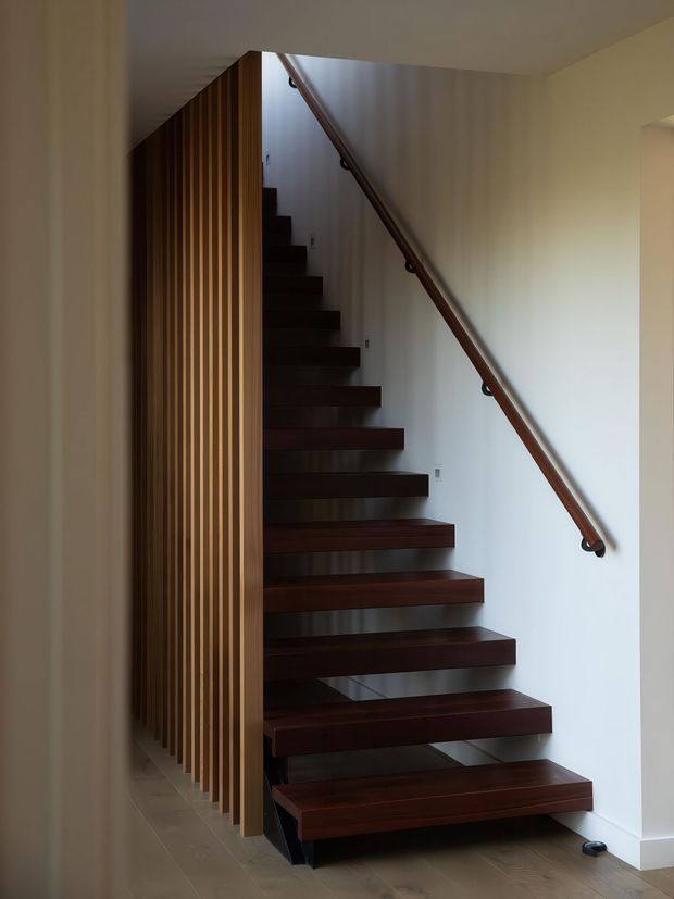 Interior view of a modern stairway with dark timber floating treads, a simple timber handrail, and a vertical timber slat screen forming the stair enclosure. Pale walls and light tiled flooring create a restrained, contemporary composition.