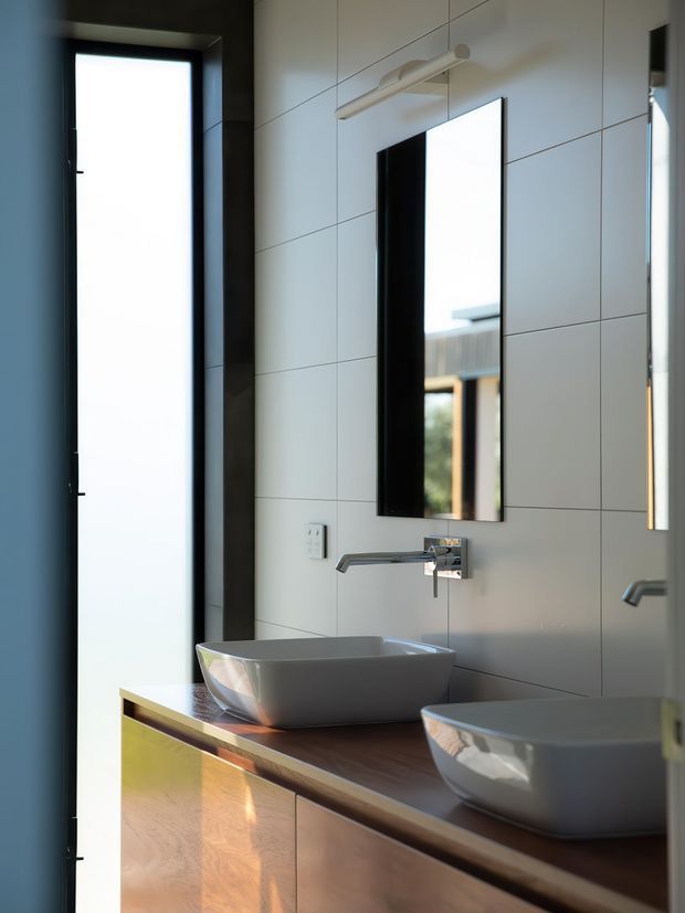 Contemporary bathroom vanity with two vessel basins on a dark timber benchtop, wall-mounted chrome faucets, large rectangular mirrors, and pale large-format tiled walls. A tall narrow window provides strong natural light, while the mirrored reflection reinforces the double-sink arrangement.