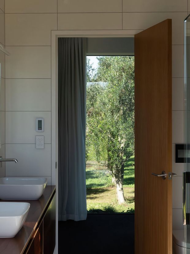 A contemporary bathroom viewed toward an open timber door leading to a leafy garden. The room features large pale wall tiles with fine dark grout lines, a dark stone or composite vanity benchtop, two white vessel basins, timber cabinetry, a wall-mounted control panel, dark curtains, and a frameless or minimally framed glass shower edge at right. Natural light and greenery outside create a calm connection to the garden.