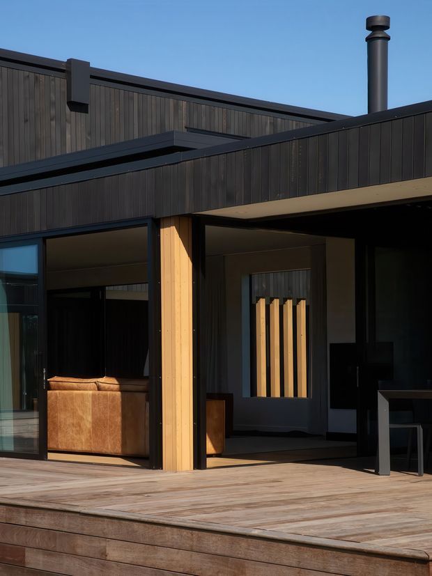 Exterior view of a modern single-storey home with dark vertical timber cladding, black-framed glazing, and a low-pitched roofline. A covered outdoor terrace with timber decking sits in front of the recessed living area, with warm timber columns and a visible cylindrical chimney. The composition highlights the building facade and deck in warm late-day light.