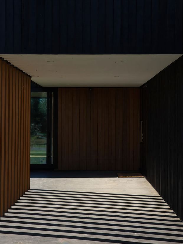 A dark, minimalist covered entryway or carport with strong linear geometry. Vertical timber or dark-stained slatted wall cladding surrounds a pale smooth ceiling and floor, while sunlight casts dramatic parallel shadows across the ground. A narrow glazed opening at the rear provides a view into landscaped greenery.