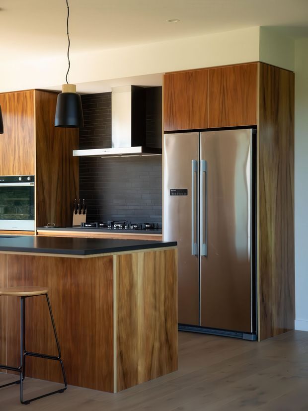 Warm modern kitchen featuring extensive rich timber cabinetry, a dark horizontal-tile splashback, black stone island benchtop, stainless-steel French-door refrigerator, integrated oven, gas cooktop, and a black pendant light. The open-plan space has timber flooring and a compact island with bar seating.