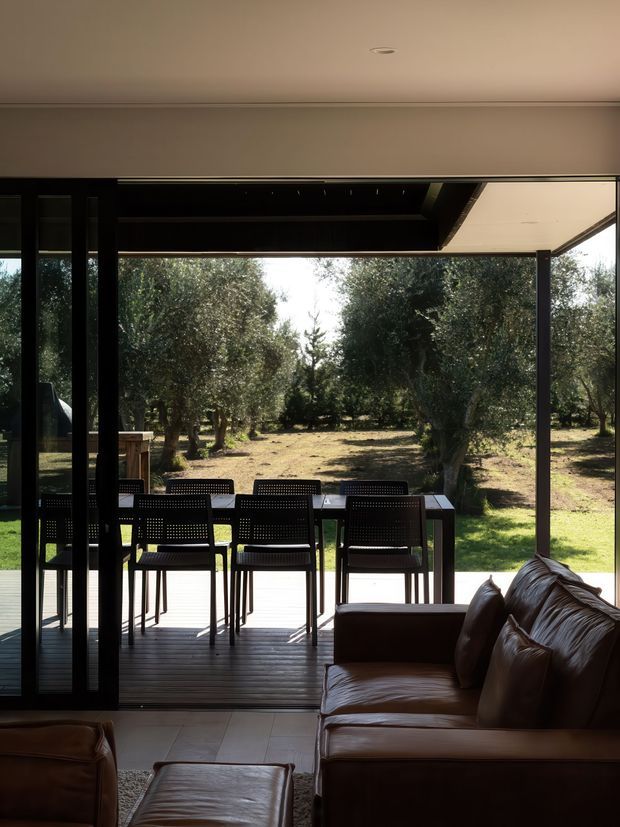Interior view of a contemporary living room opening onto a covered timber deck and outdoor dining area. Large black-framed sliding glass doors create broad views toward a wooded, landscaped garden. The room contains a dark brown leather sofa and ottoman, while the deck has a long dining table and perforated-back chairs. A deep covered roof overhang and dark structural framing define the indoor-outdoor connection.